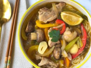 A bowl of chicken sop with limes, chili peppers, noodles, bell peppers, garlic and stock.