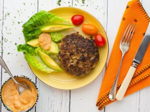 A plate with a burger and salad on it, with a dip on the side with an orange napkin and knife and fork.