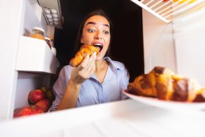 A female putting acroissant into her mouth with her head in the fridge looking for more food.