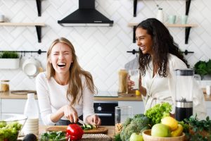 Two females laughing out in the kitchen, while chopping vegetables.