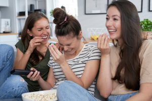 Three girlfriends laughing on the sofa eating popcorn, presumably watching a comedy.