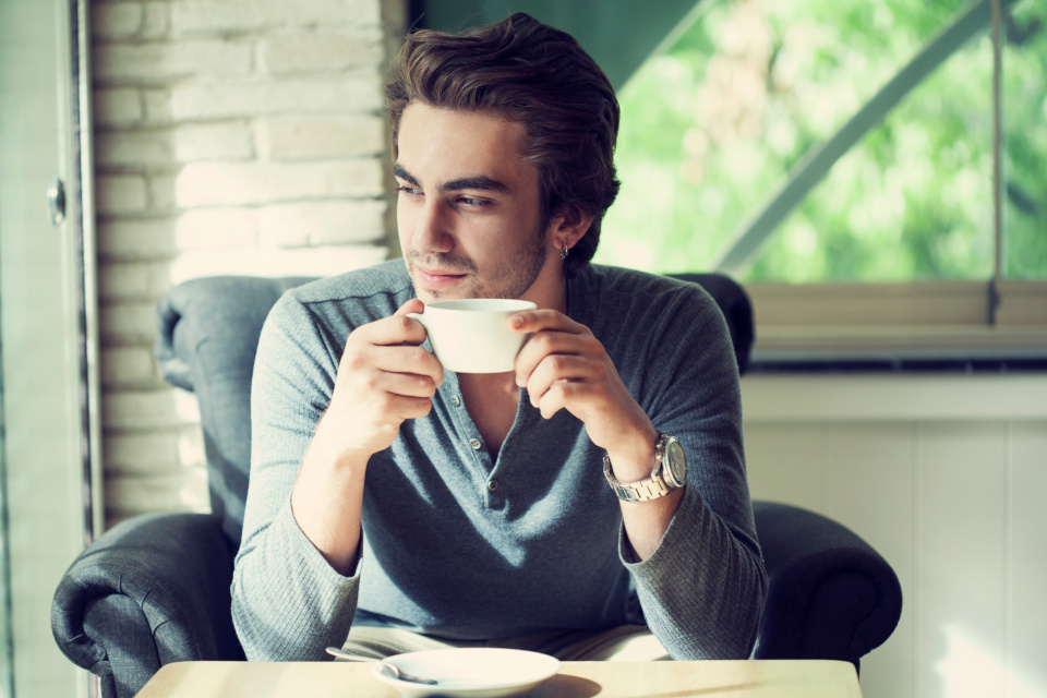 Dark haired man sat alone in a grey upholstered chair drinking tea, looking calm with a pleasant look on his face