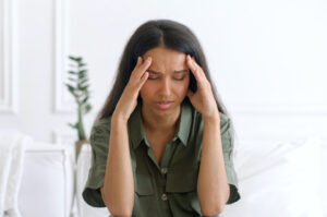 Stressed young indian woman touching head and feels dizzy, suffering from headache and migraine pain, sits on the sofa at home with eyes closed, tired and exhausted mixed-race lady feels bad