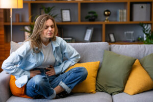 A young female sitting on a sofa clutching her stomach, indicating stomach or period pain.