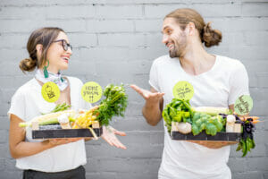 Young vegetarian couple standing with boxes full of fresh vegetables outdoors on the gray wall background