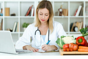 Female vegan and vegetarian nutritionist working at desk and writing medical records over fresh fruit in the consultation.