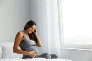 Female, pregnant, sat on her bed, smiling and holding her tummy. 