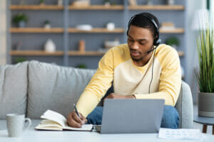 Young guy attending online training or webinar from home, sitting on couch in front of coffee table with laptop, using headset, taking notes, copy space. E-education, online class, training concept